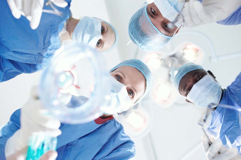 four people in scrubs and medical masks look down toward the camera lens. One woman holds a tube to deliver anesthesia.