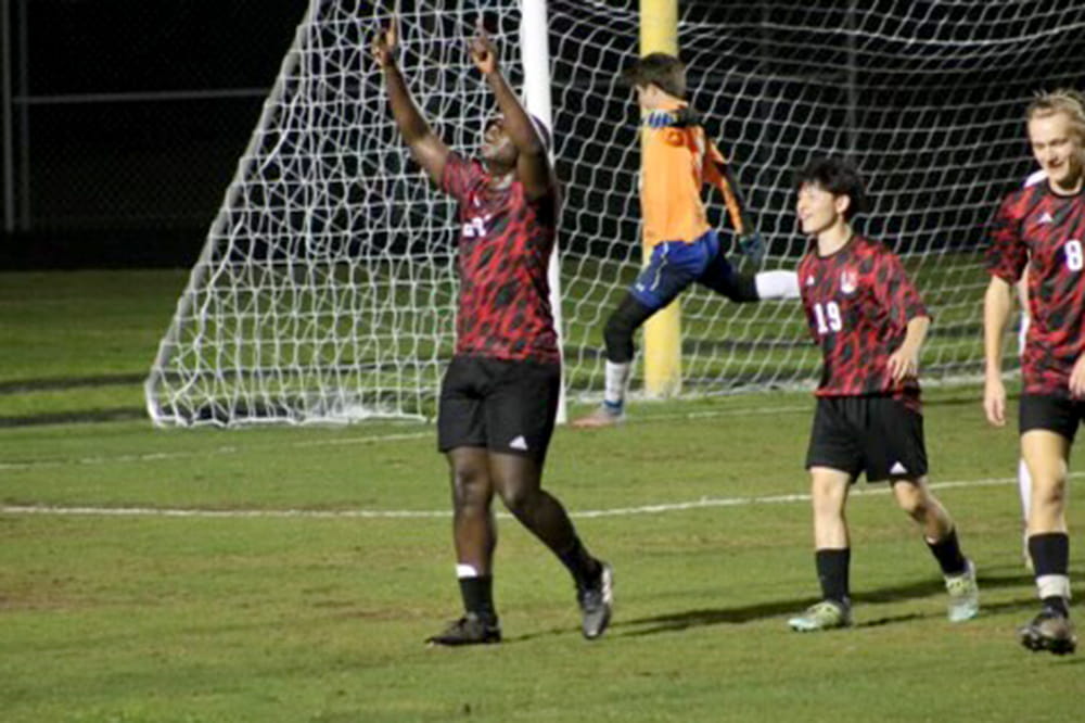 Young man in red and black uniform raises arms in celebration. Three other soccer players are behind him.