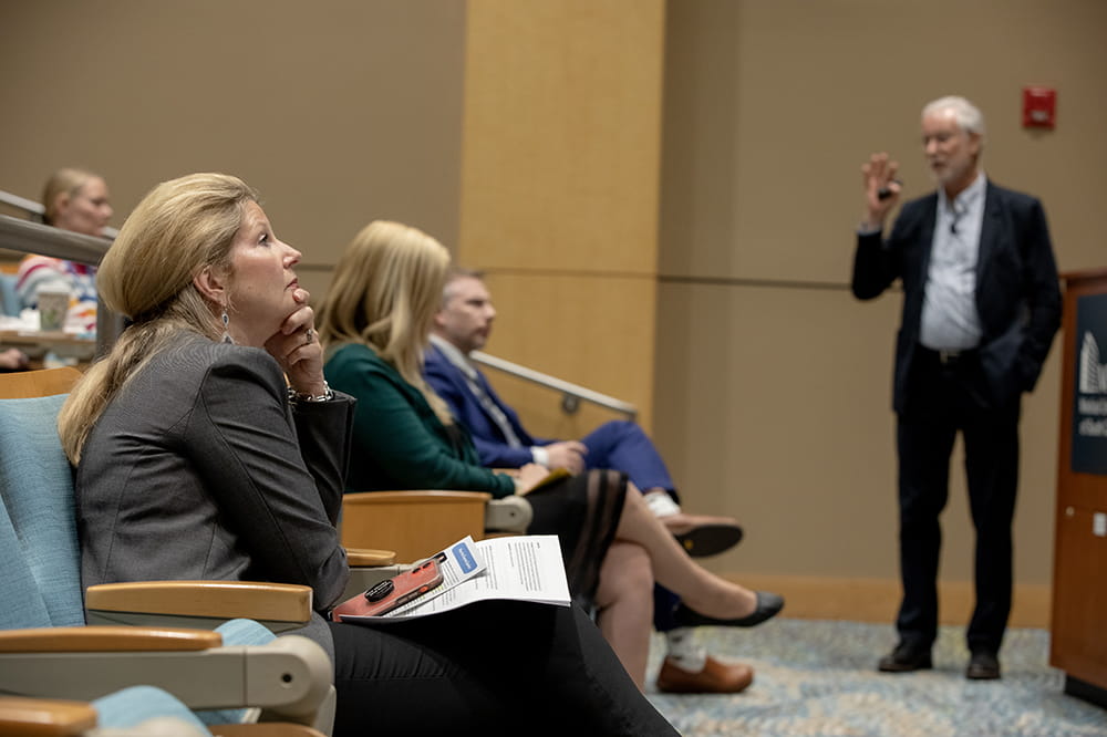 Woman with blonde hair leans an her arm as she looks intently forward. A man is standing and speaking. He is wearing a suit.