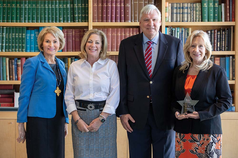 Three women and one man smile while standing in front of a bookshelf. The woman on the far right is holding an award.
