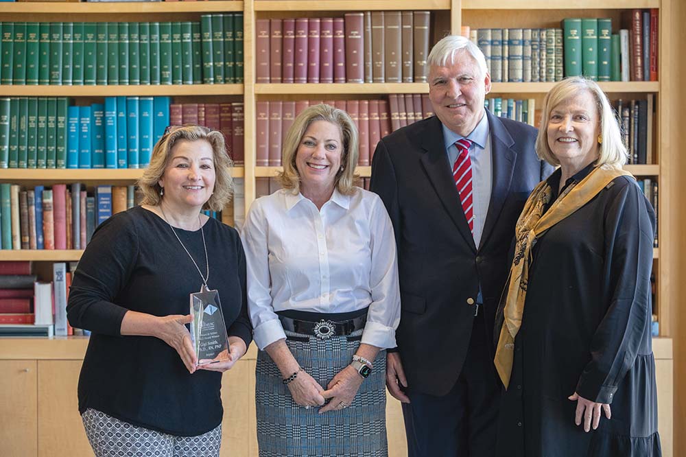 Three women and one man smile in front of a bookshelf. One of the women is holding an award.
