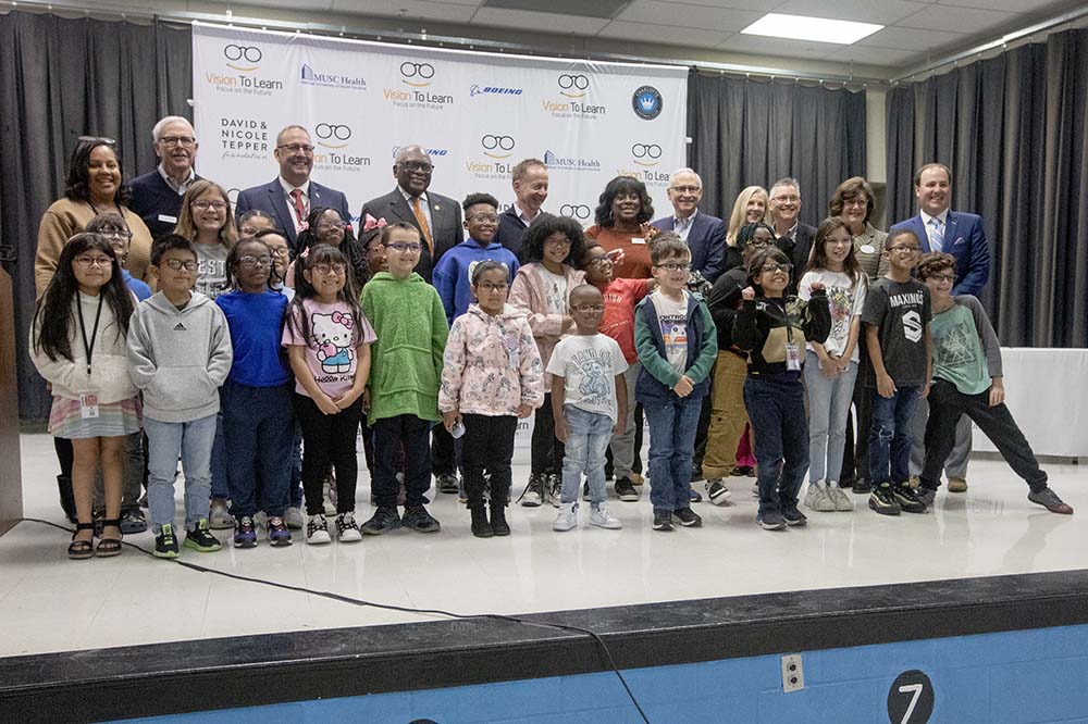 Children and adults stand on stage smiling. The kids are wearing new eyeglasses.