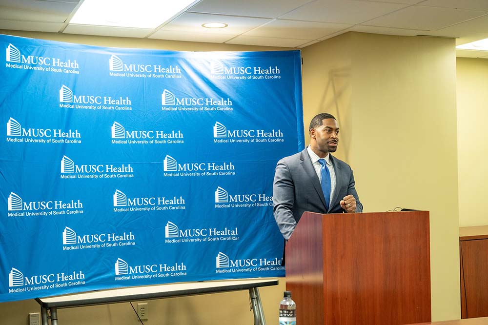 Man in a suit standing at a podium. There is a sign saying MUSC Health behind him.