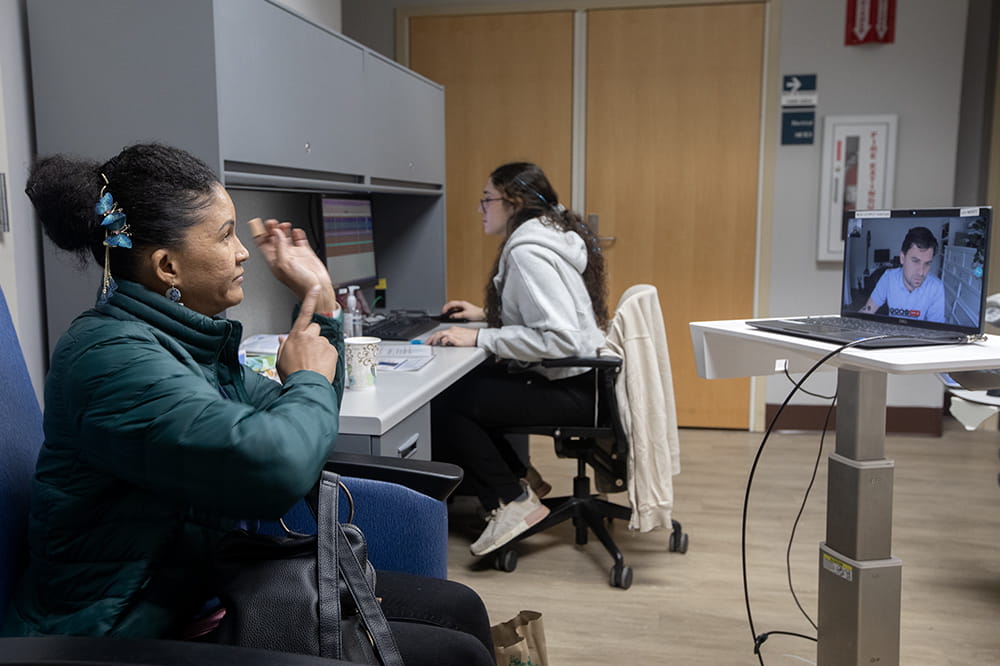 Woman in a chair point to her left hand. She's looking at a screen where she sees a man.