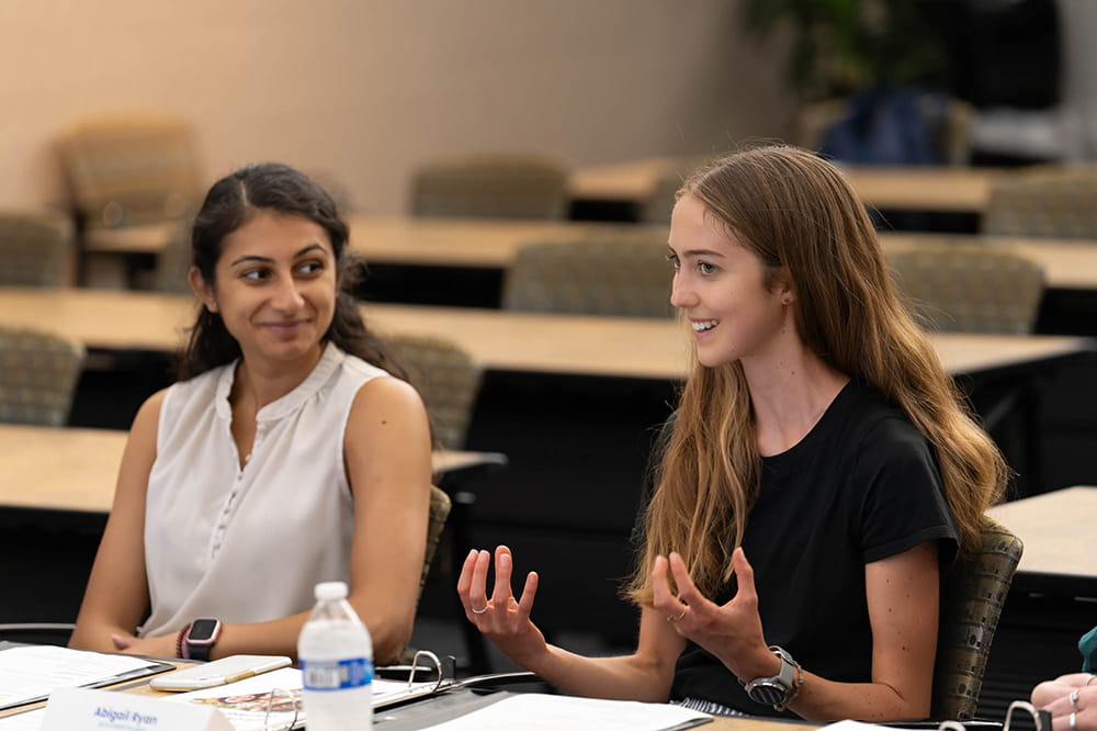 two young women sit side by side at a table in a large meeting room. One looks at the other with a smile as the other talks and gestures with her hands