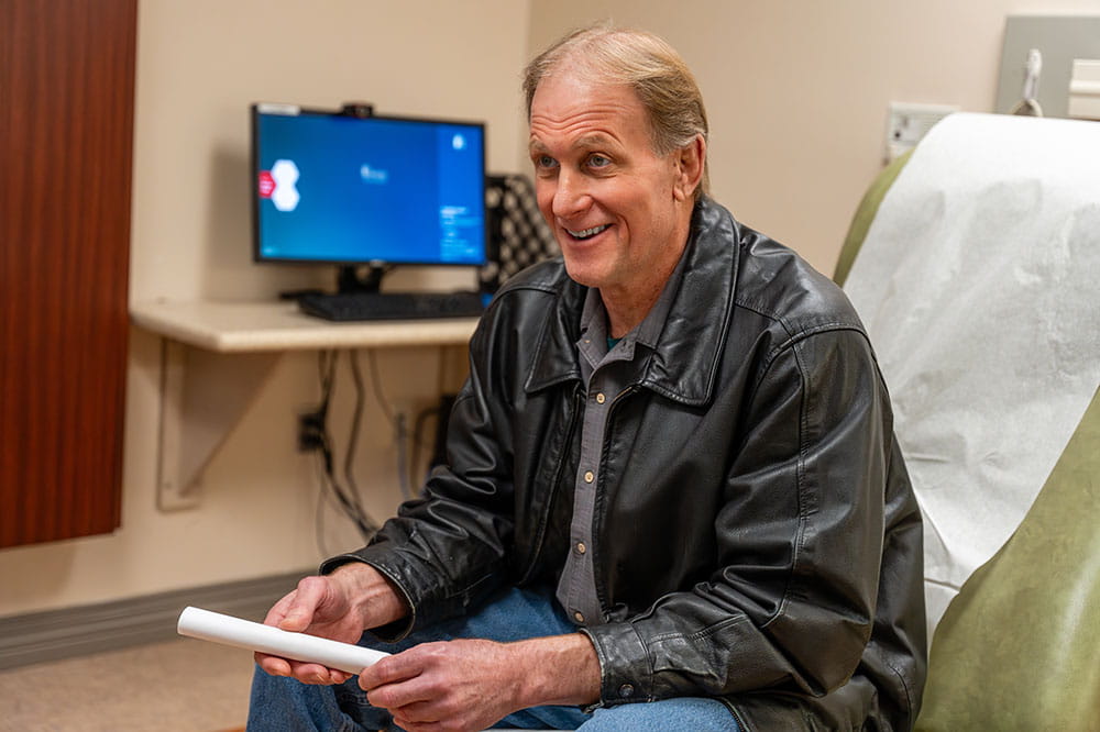 a man in jeans and loose black leather jacket leans forward, smiling broadly, while sitting on an exam chair in an exam room