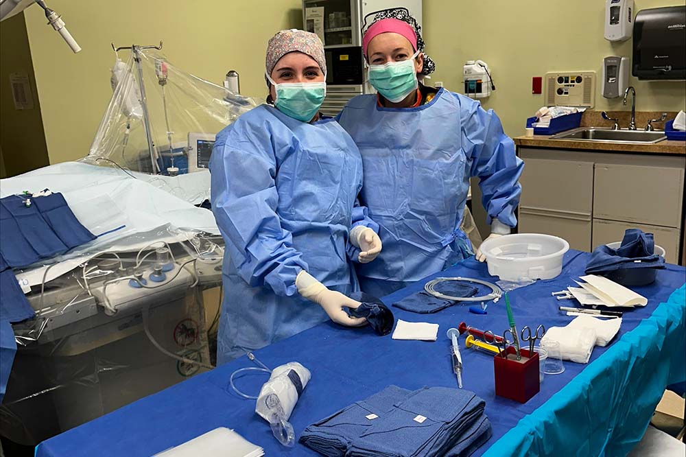 Two women in scrubs and masks smile while standing in a cath lab.