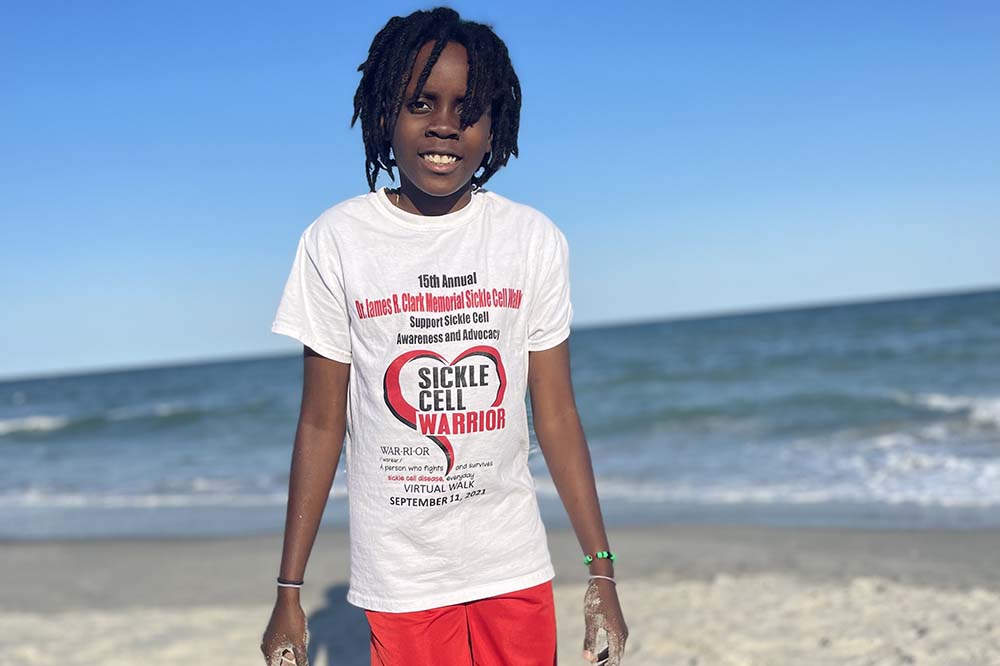 A young man stands in front of the ocean smiling. He's wearing a t-shirt that says sickle cell warrior.