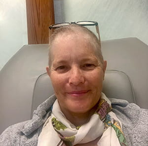 a woman sitting in a infusion treatment chair smiles at the camera, her glasses perched atop her head and a scarf swaddling her neck. Her hair is less than an inch long