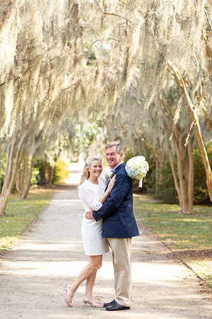 a bride and groom pose in a park with an allee of crepe myrtles dripping with Spanish moss behind them, the bride dressed in a knee length white dress and the groom in blazer and khakis