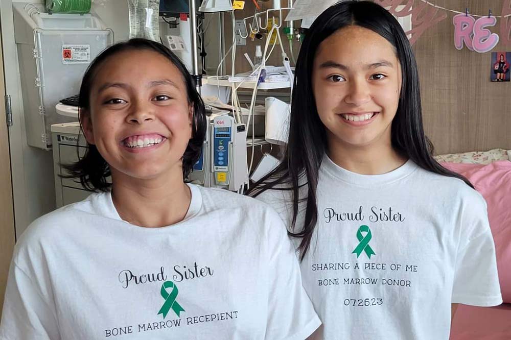 Two girls with dark hair. They are wearing white t-shirts that say proud sister and have a green ribbon.