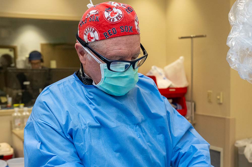 Man with red cap that says Red Sox, eyeglasses and a medical mask looks down.