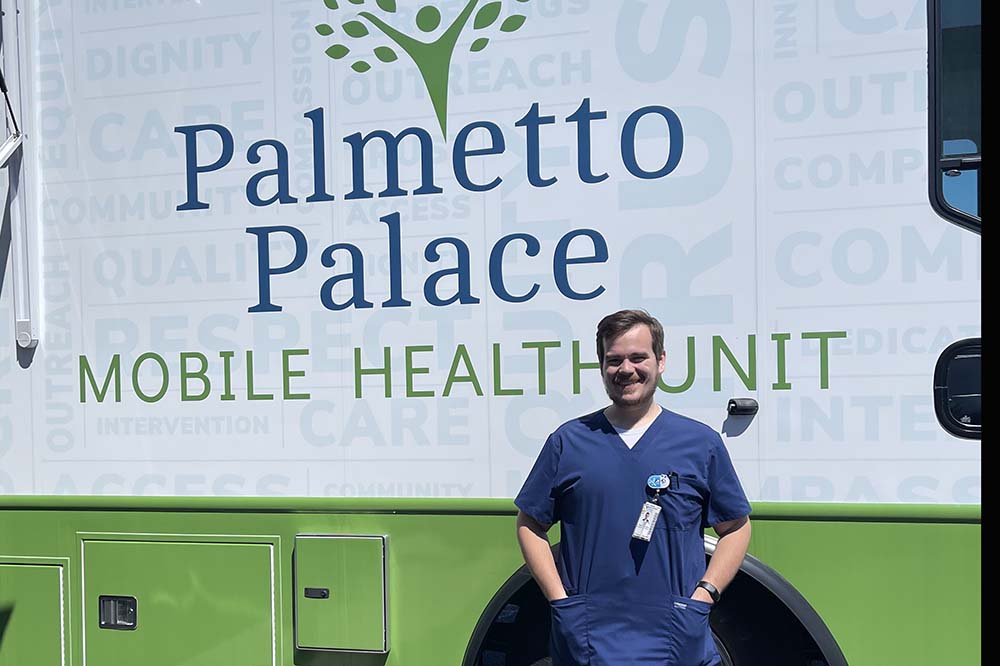 Man in blue scrubs stands in front of truck with the words Palmetto Palace Mobile Health Unit.