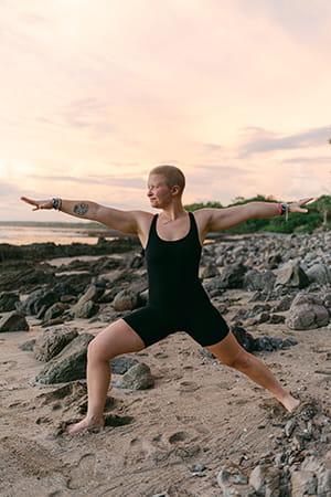 a young nearly bald woman in black yoga shorts and top does a warrior pose on the beach