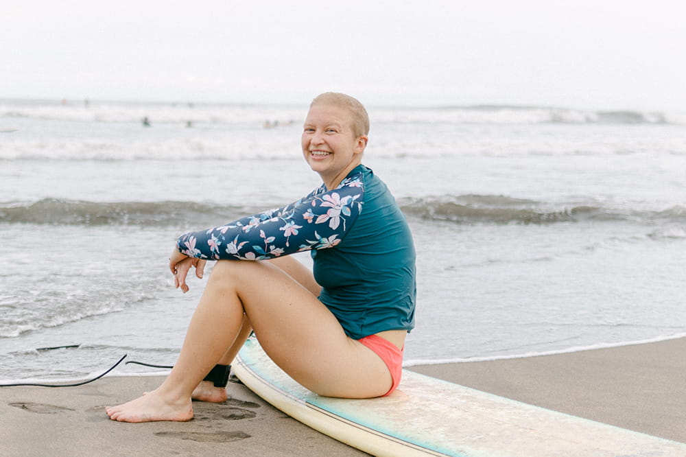 a young nearly bald woman in a green rash guard sits on a surfboard at the water's edge