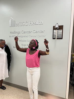 a woman in pink tank top and white jeans raises her arms in victory while in front of a bell rung by patients after they finish treatment