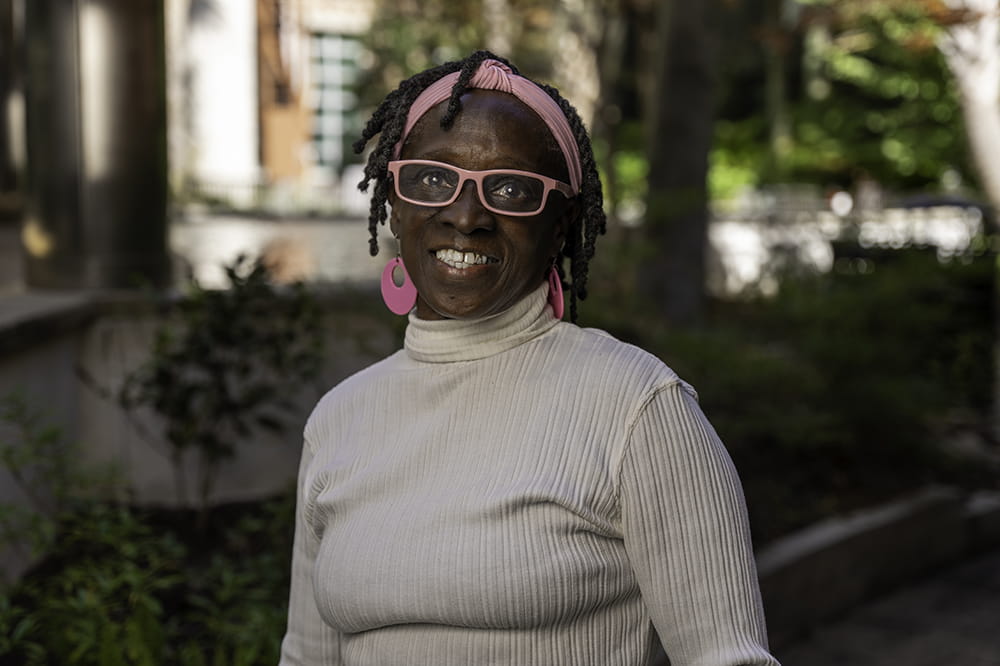 a woman in bright pink headband, glasses and earring smiles in a garden setting