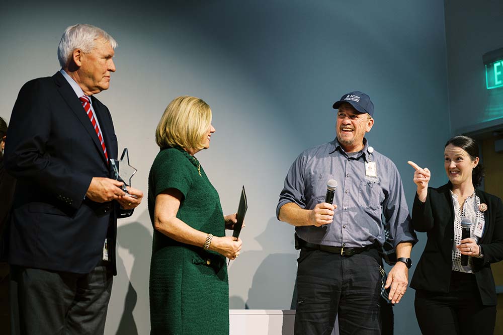 Two women and two men are on stage. The man on the right is wearing working clothes and holding a microphone.