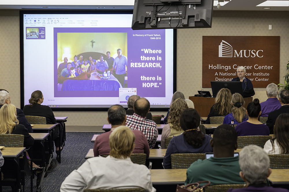 a short older woman in a purple shirt stands at a podium in front of a large group with an image on the screen behind her of her late husband and the family