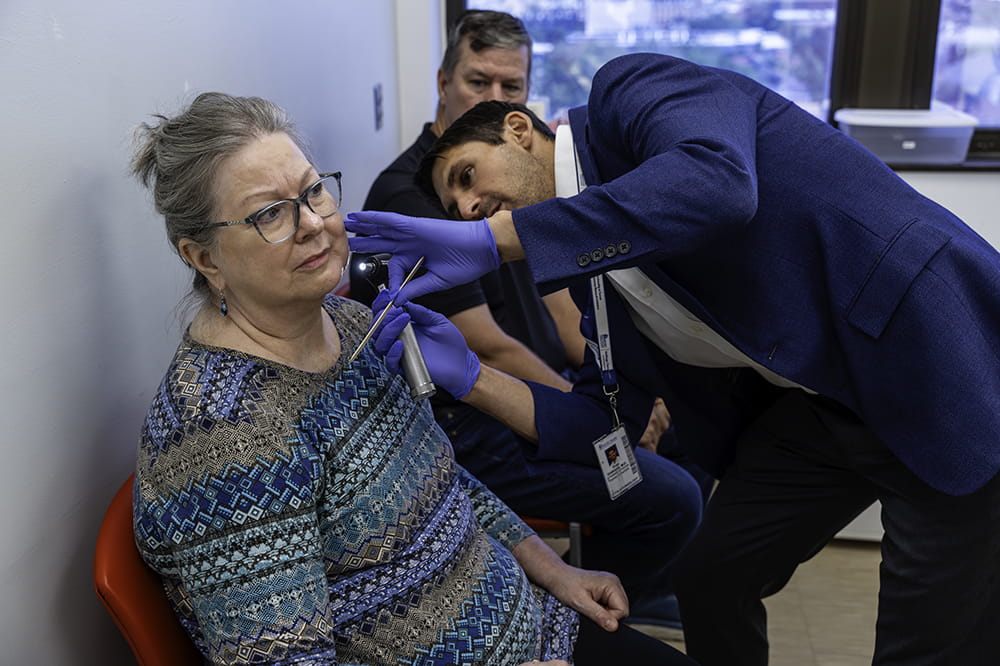 a patient sits in a chair while a doctor leans over to the side to look at the side of her face