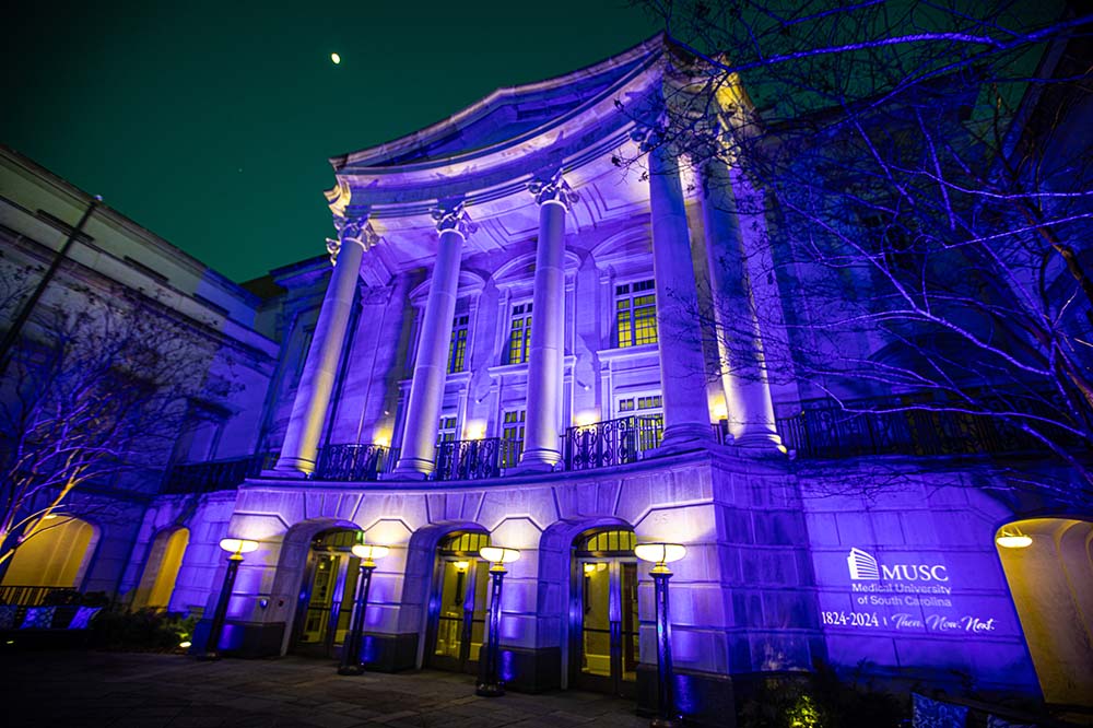 Outside of a building that is covered in blue light and has the words Medical University of South Carolina 1824-2024, then, now, next on the lower right corner.