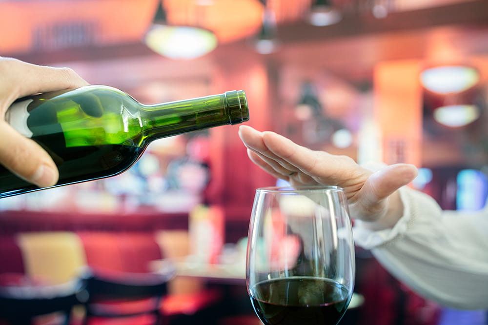 as someone attempts to pour more red wine into a wine glass, the drinker places her hand over the glass indicating she doesn't want any more