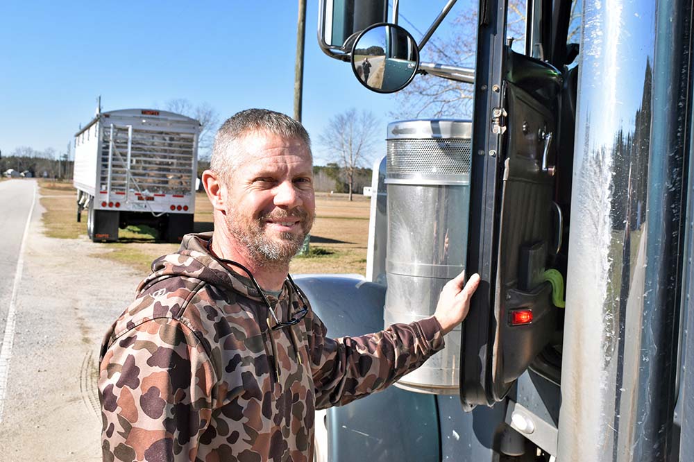 A man stands with his hand on the door of a large truck. He has gray hair and a beard and is smiling.