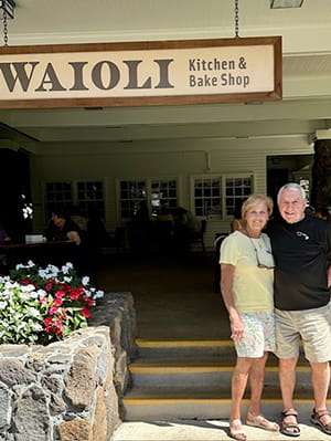 a couple in shorts and tee shirts poses in front of a shop called Waioli Kitchen and Bake Shop
