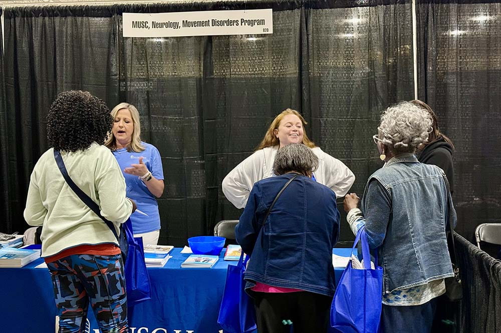 Two women stand behind a table while three other women talk to then. There's a sign that says MUSC movement disorders.