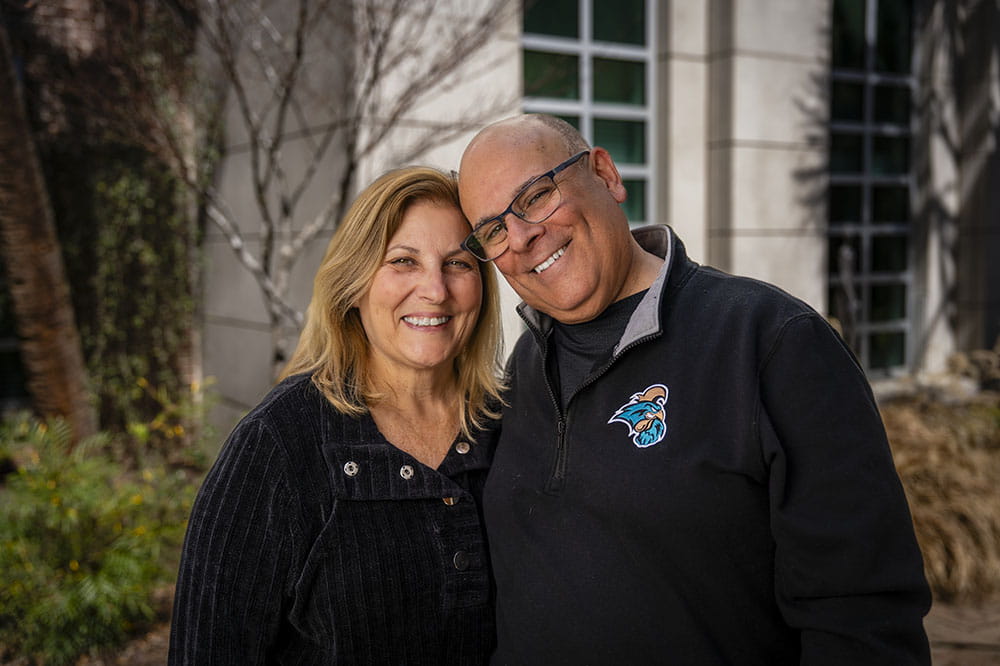 A man and woman pose in a garden setting, the man tilting his head to touch the woman's head