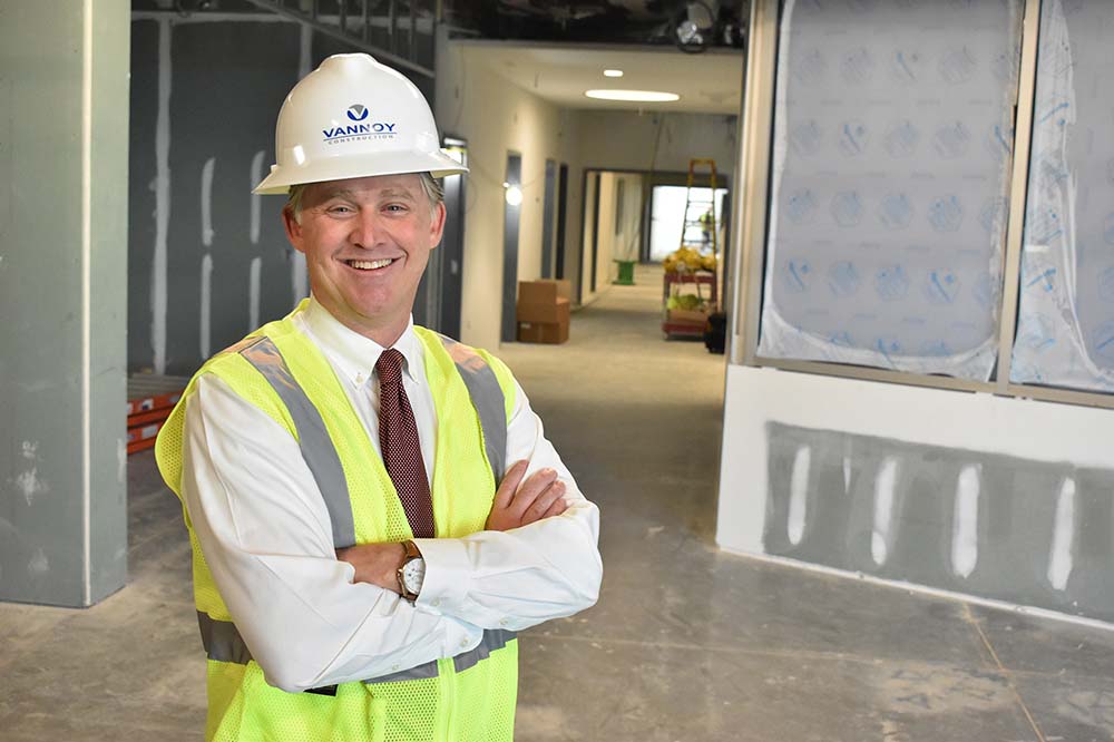 Man in a shirt and tie wears a bright yellow vest and a construction helmet. He is standing in a construction site.