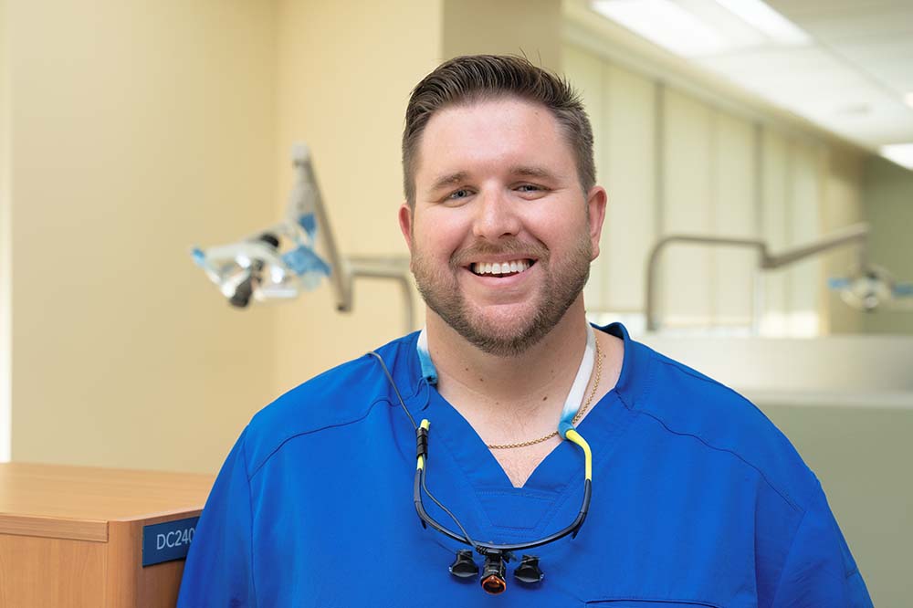 Headshot of smiling bearded man. He is wearing blue scrubs and has a dental instrument hanging from his neck.