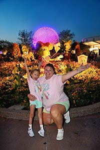 A woman with a girl raising their arms in front of a purple globe at Disney World.