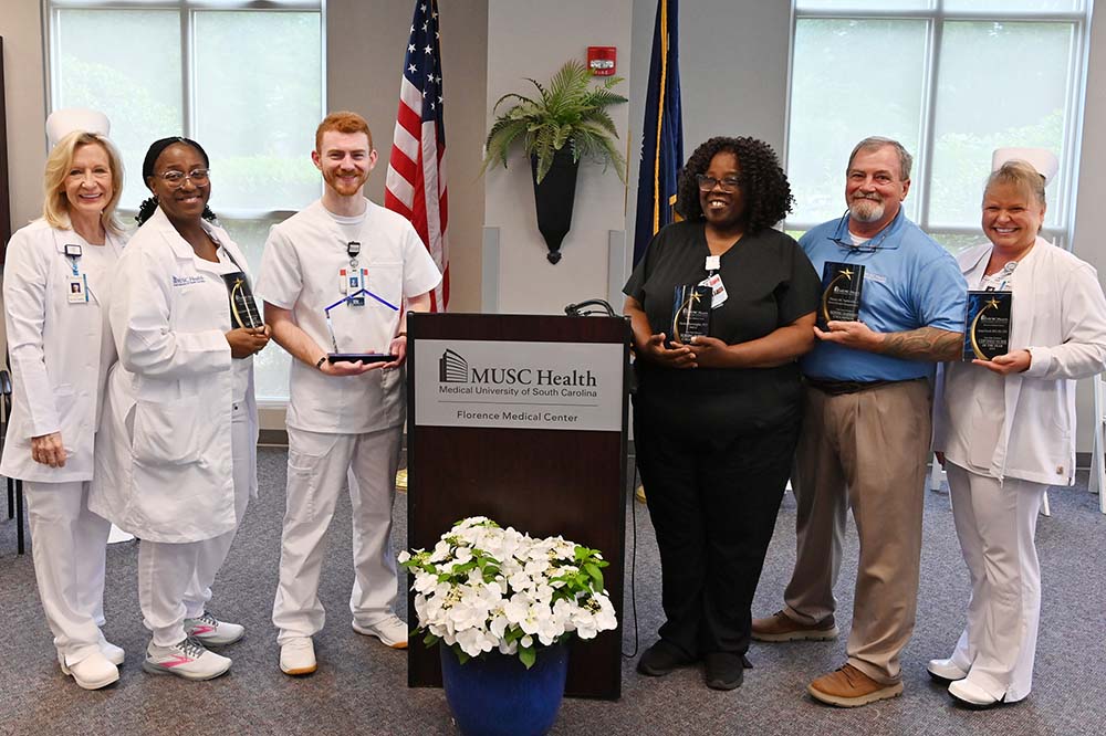 Six people stand beside a podium dressed in hospital scrubs. There is a flowering plant in front of the podium. the people are holding awards. 