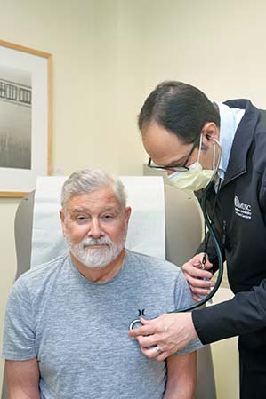 A man with gray hair and a beard is seated as a soctor examines him. The patient is sitting in an exam chair. The doctor is wearing a surgical mask and dark clothing.