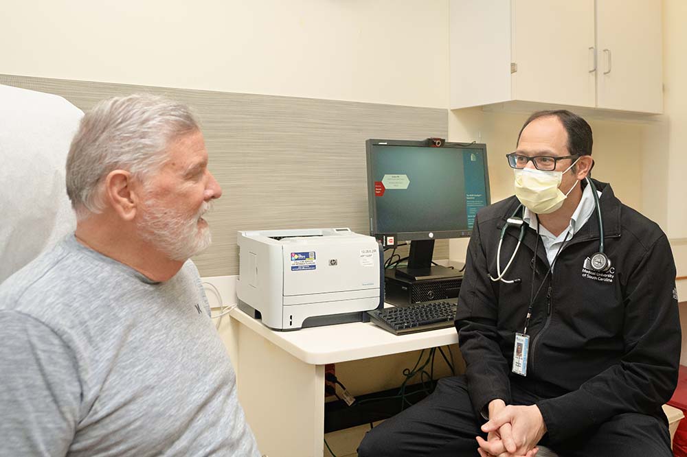 A man with gray hair and a beard wearing a t-shirt talks to another man who is wearing a surgical mask, a stethoscope and dark clothing.