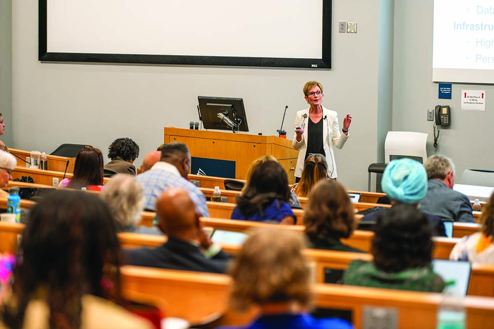A woman with short, light hair and glasses stands in the front of an auditorium
