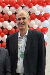 Man wearing an open collared shirt and a jacket smiles standing in front of red and white balloons.