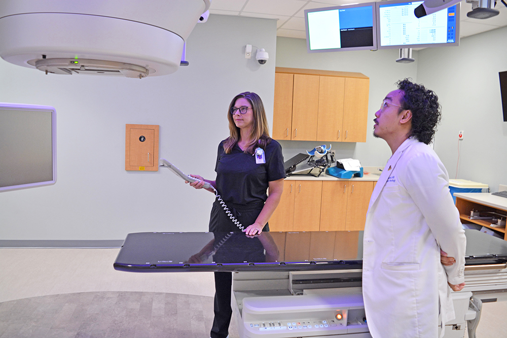 a doctor in white coat and a radiation therapist stand in the radiation oncology room and look up at the machine