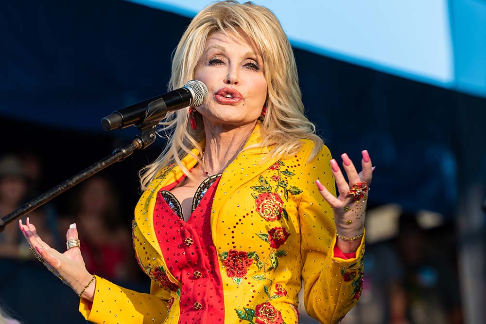 A blonde woman wearing a yellow jacket gestures with her hands.