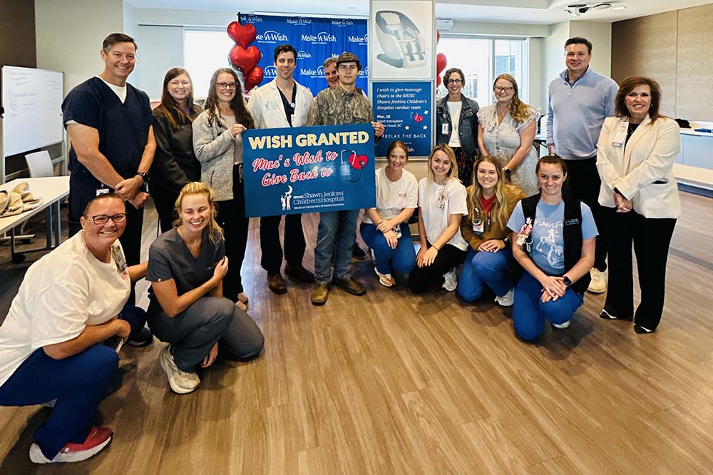 A young man wearing a camouflage cap and jacket holds a sign that says Wish granted. Mac's wish to give back to MUSC Shawn Jenkins Children's Hospital.