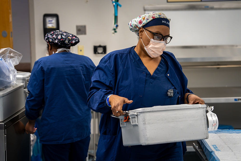 a woman carries a metal case full of OR supplies while another nurse works behind her