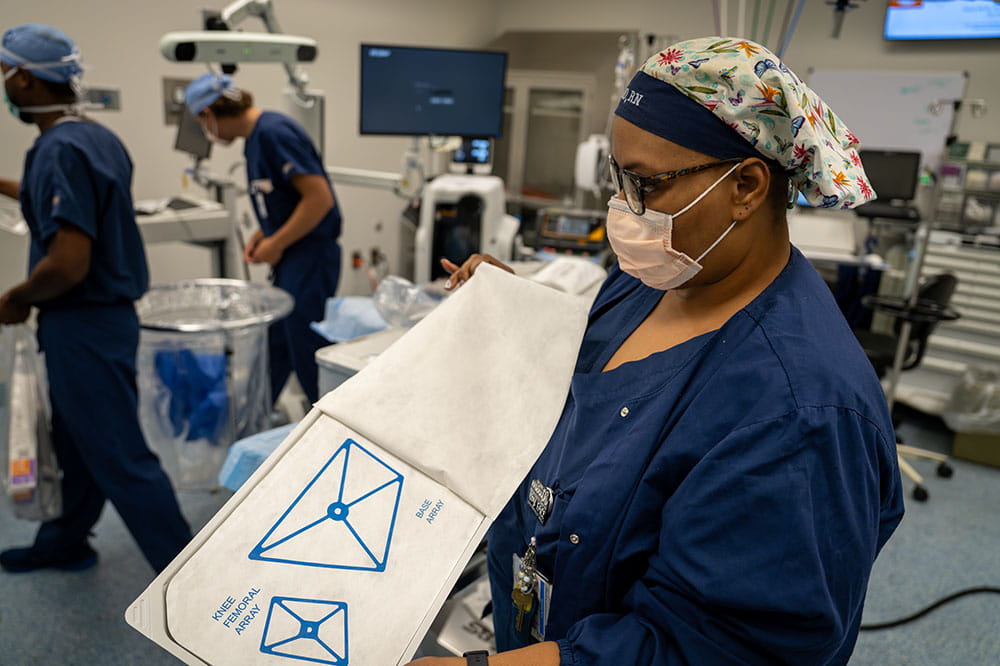 a woman opens a sealed package of operating room materials while two people in the background work on setting up other things