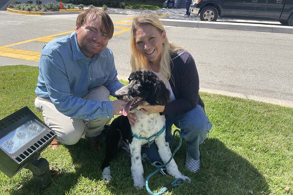 A young man and a woman crouch with a black and white dog. All three look happy. They are outside on grass.